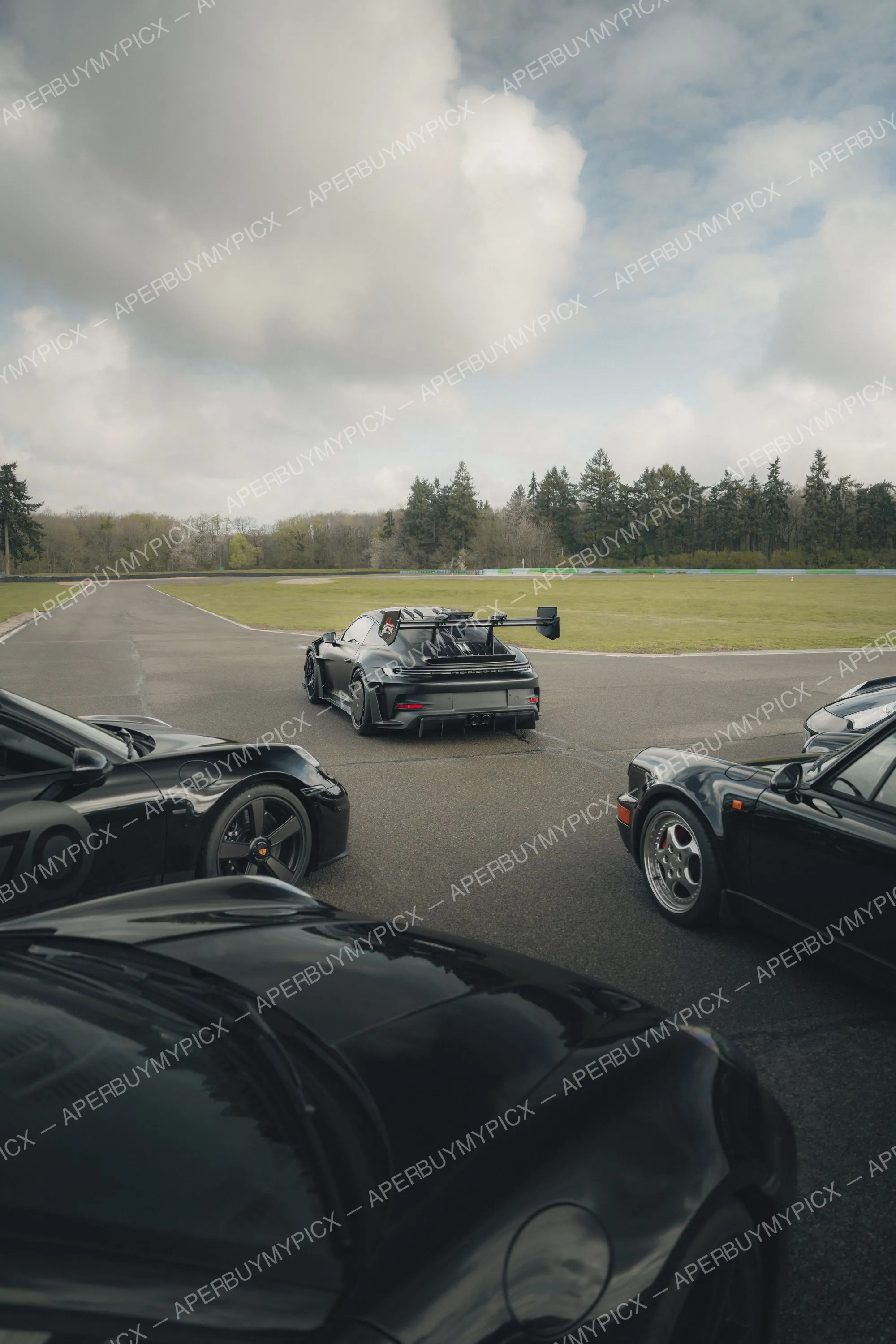 Porsche 911 GT3 RS noire avec aileron arrière sur un circuit, entourée de plusieurs voitures de sport noires dont une Porsche 959 classique, sous un ciel nuageux bordé de forêts.