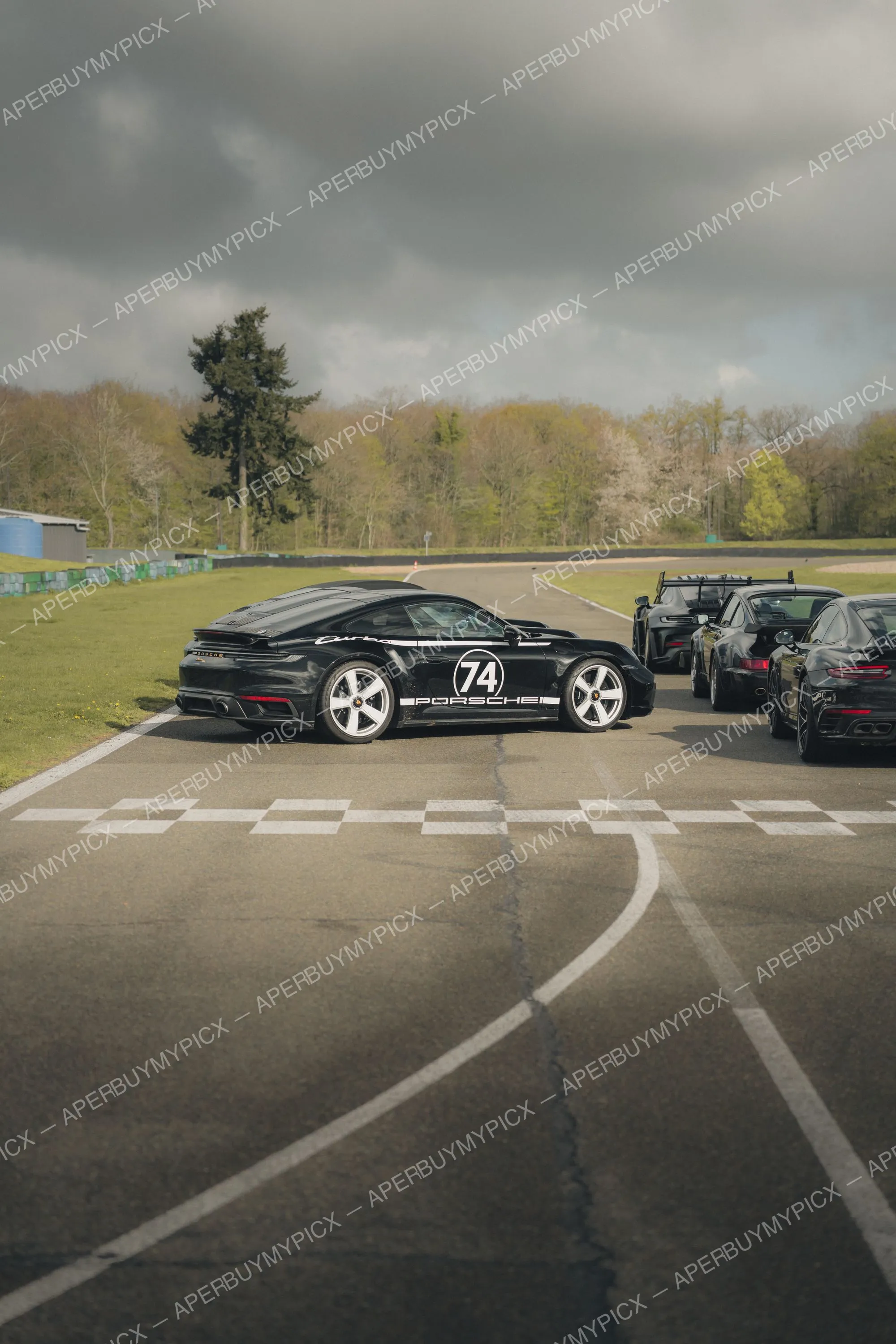 Porsche 911 Turbo noire avec livrée numéro 74 garée sur la ligne de départ d'un circuit, suivie d'autres Porsche noires, sous un ciel nuageux et des arbres en arrière-plan.