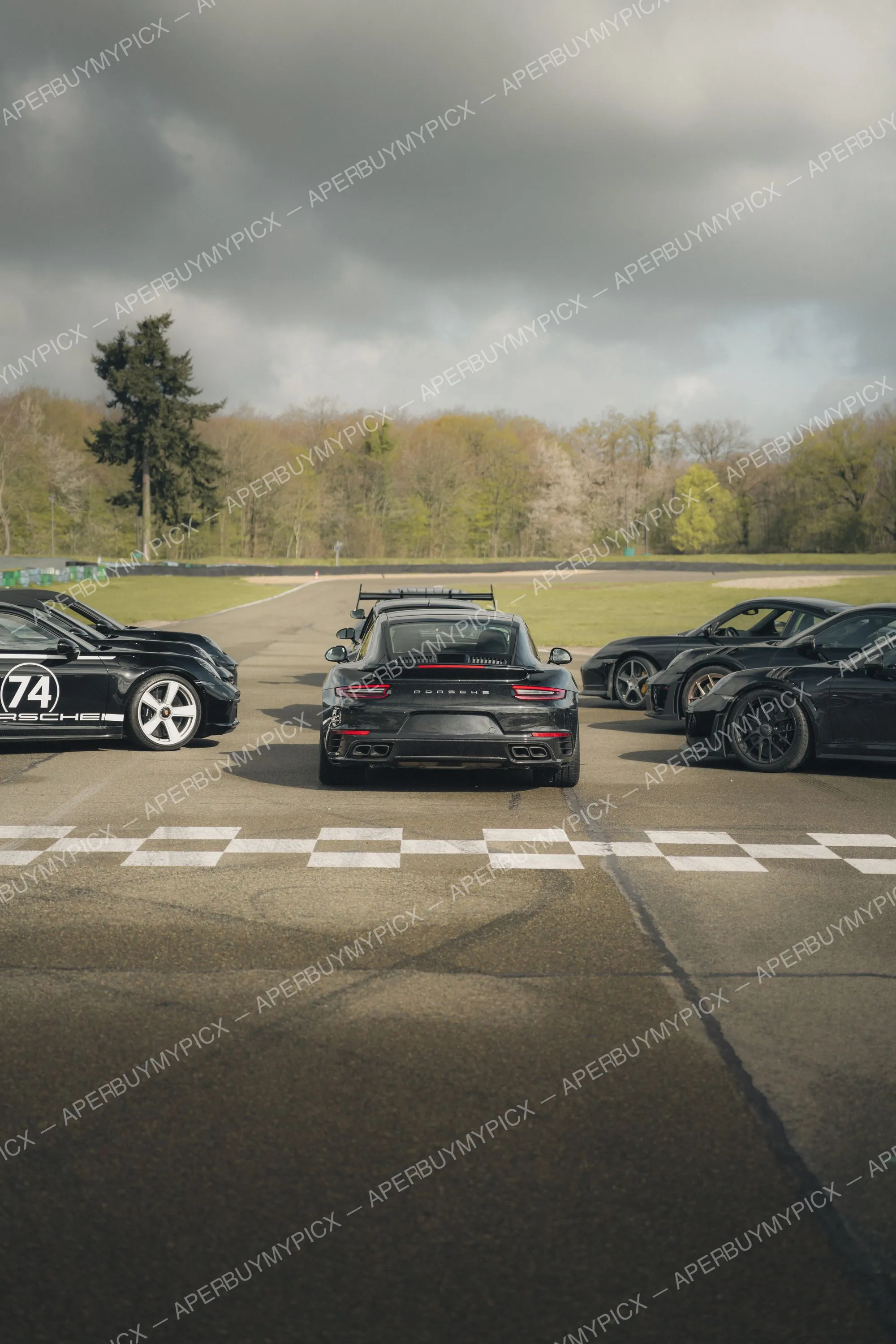 Trois Porsche 911 noires alignées sur un circuit sous un ciel nuageux, vue arrière d'une Porsche Turbo au centre près de la ligne de départ à damier