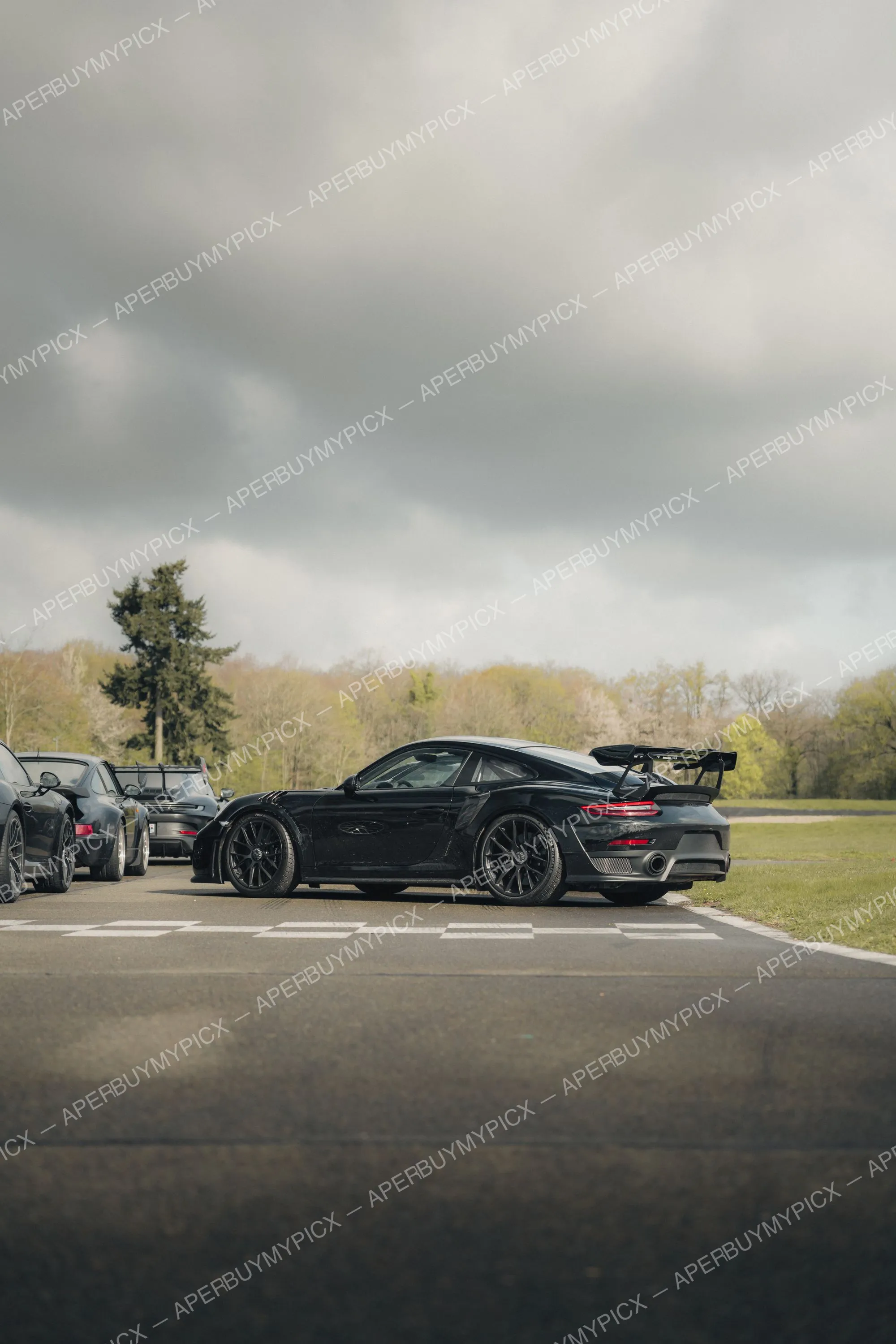 Porsche 911 GT2 RS noire avec grand aileron arrière, vue de profil trois-quarts arrière, garée sur un circuit avec des vibreurs, d'autres Porsche à gauche et une forêt sous un ciel nuageux.