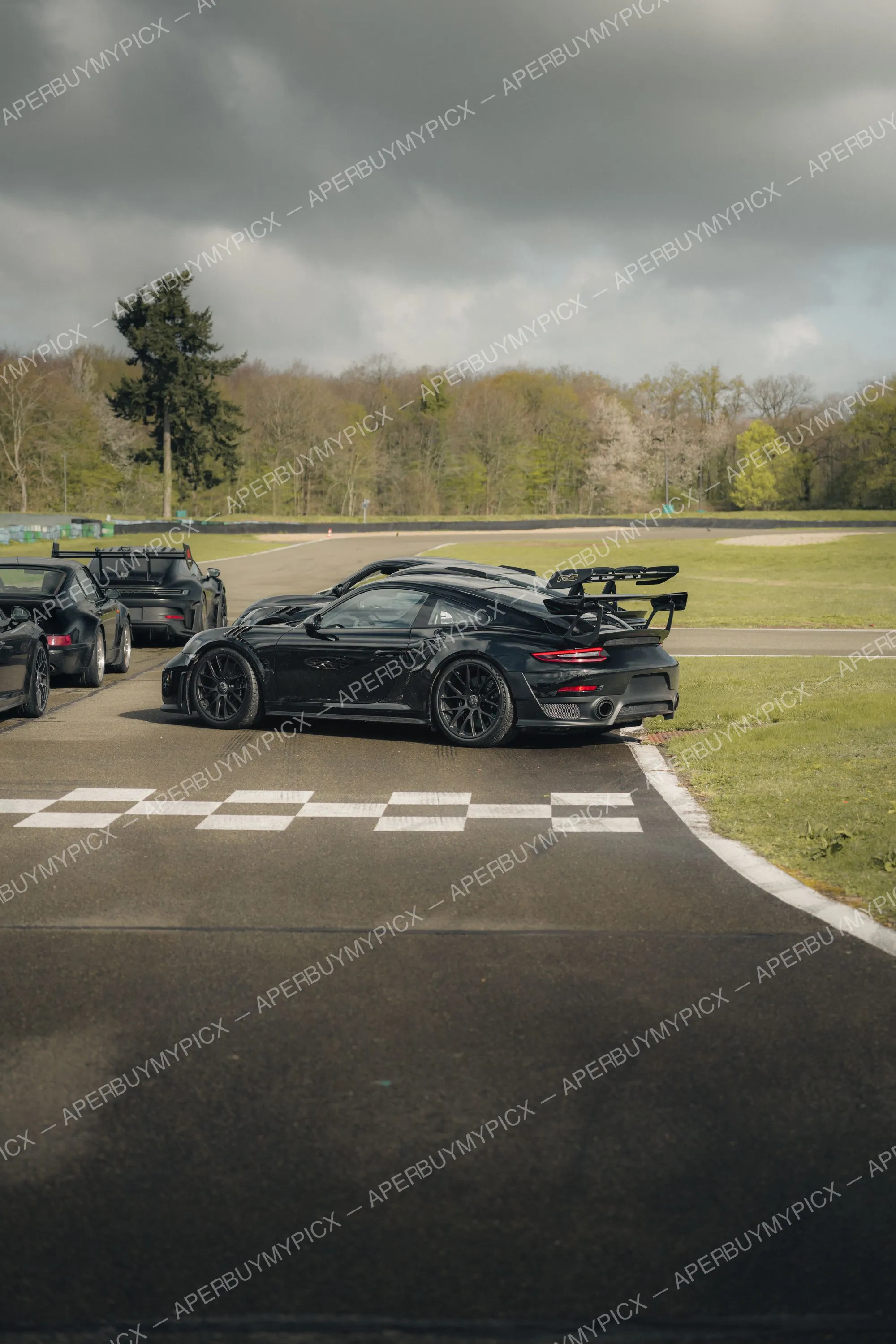 Porsche 911 GT2 RS noire sur la piste d'un circuit, aile arrière fixe, jantes noires, alignée derrière d'autres Porsche noires près de la ligne à damier sous un ciel nuageux.