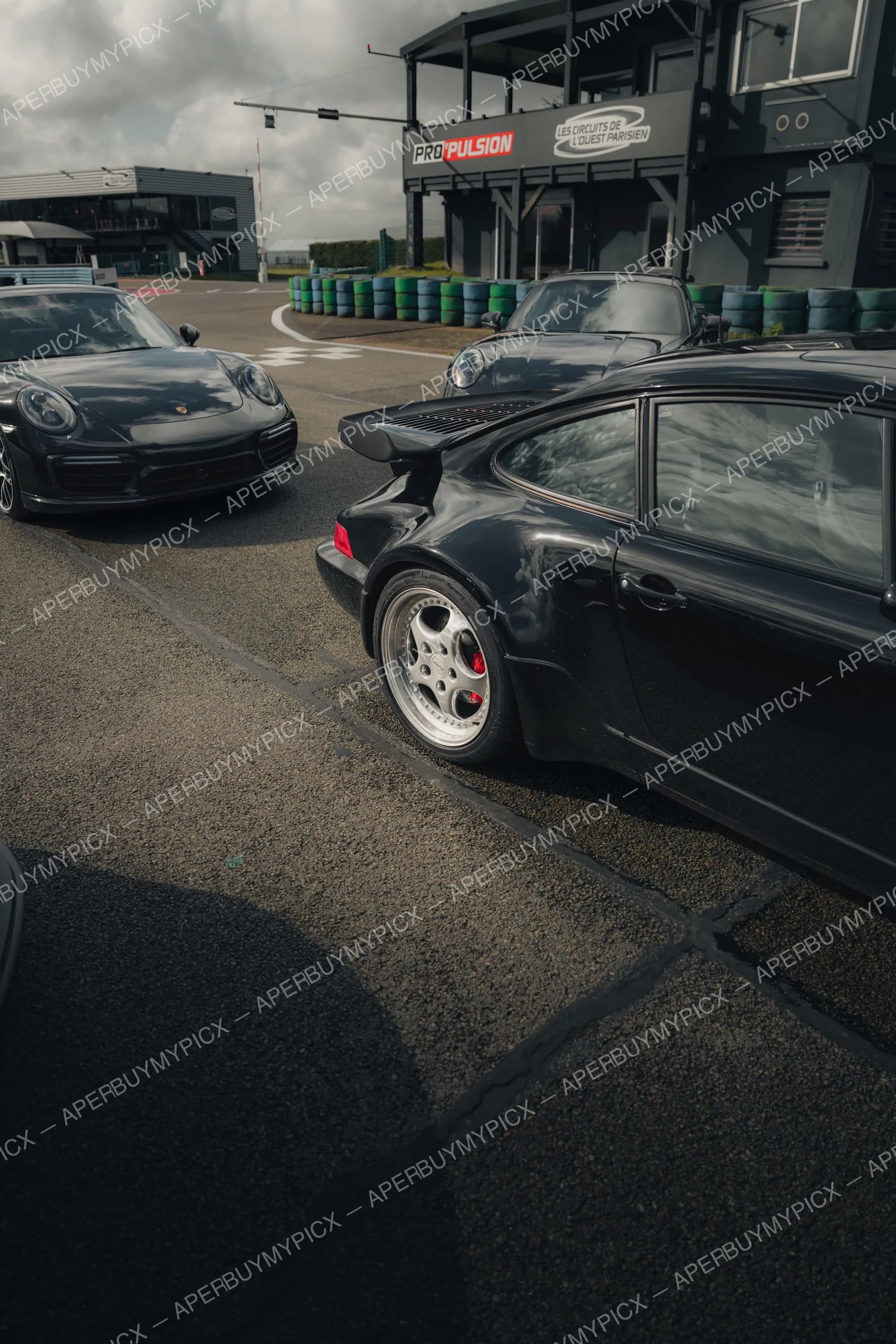Trois Porsche 911 noires stationnées sur un circuit automobile, avec la 964 Turbo à aileron au premier plan, devant le bâtiment Propulsion des Circuits de l'Ouest Parisien sous un ciel nuageux.