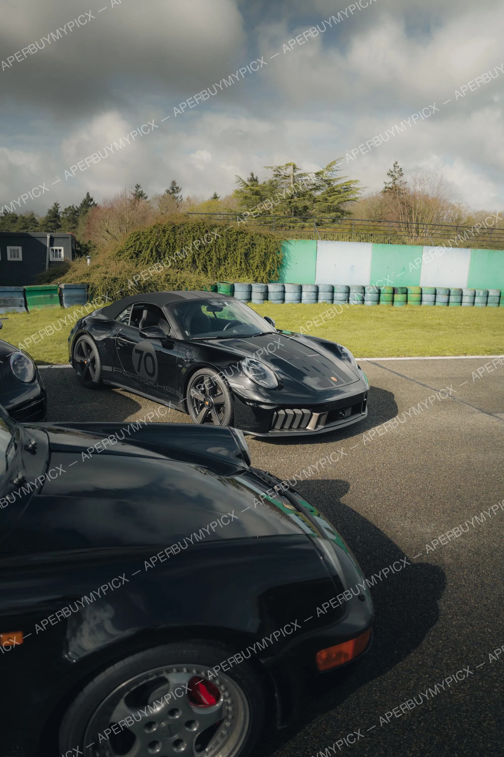 Porsche 911 Speedster noire portant le numéro 70 stationnée sur un circuit, à côté d'une autre Porsche noire au premier plan, avec pneus de protection verts, mur peint et arbres sous un ciel nuageux.