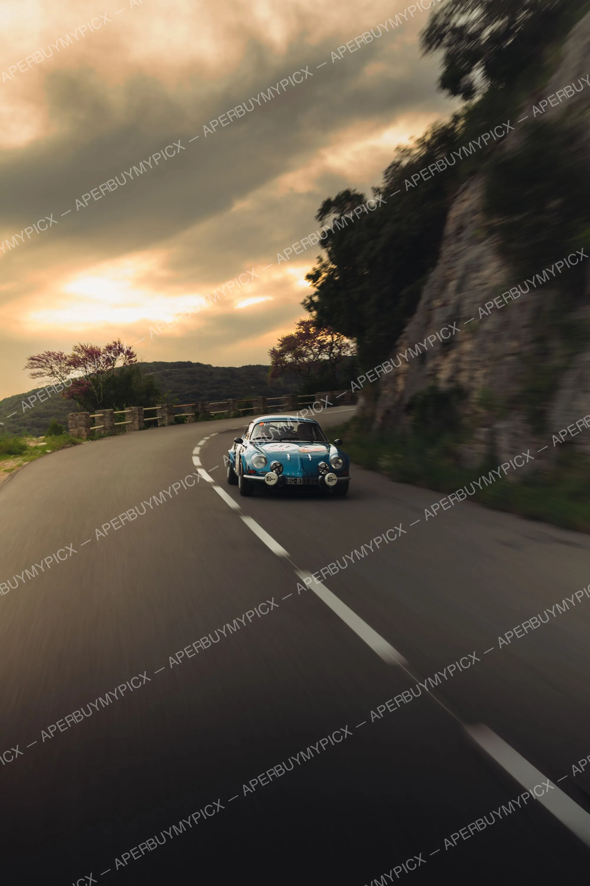 Alpine A110 bleue numéro 211 roulant sur une route de montagne sinueuse au coucher du soleil, falaise rocheuse à droite et ciel nuageux orangé