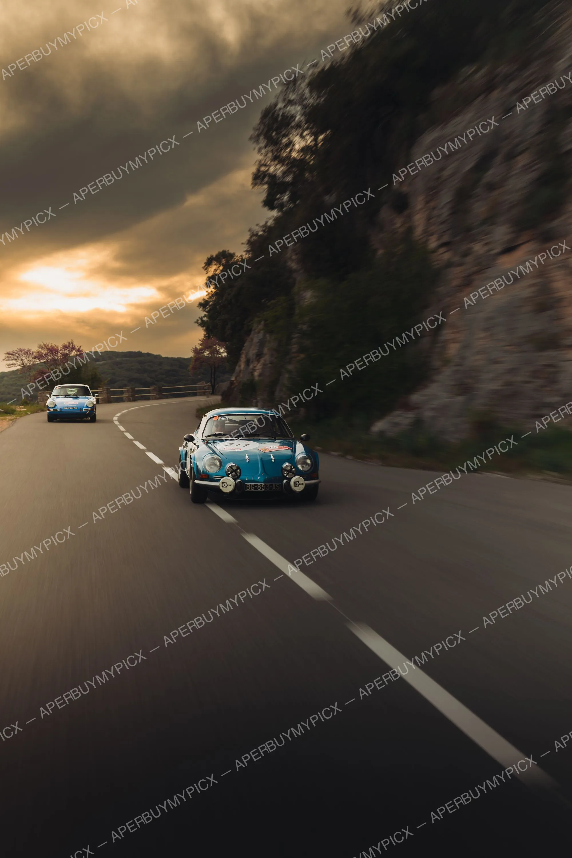 Alpine A110 bleue numéro 211 roulant sur une route de montagne sinueuse au coucher du soleil, suivie d'une Porsche 911 bleue, lors du Tour Auto 2025.