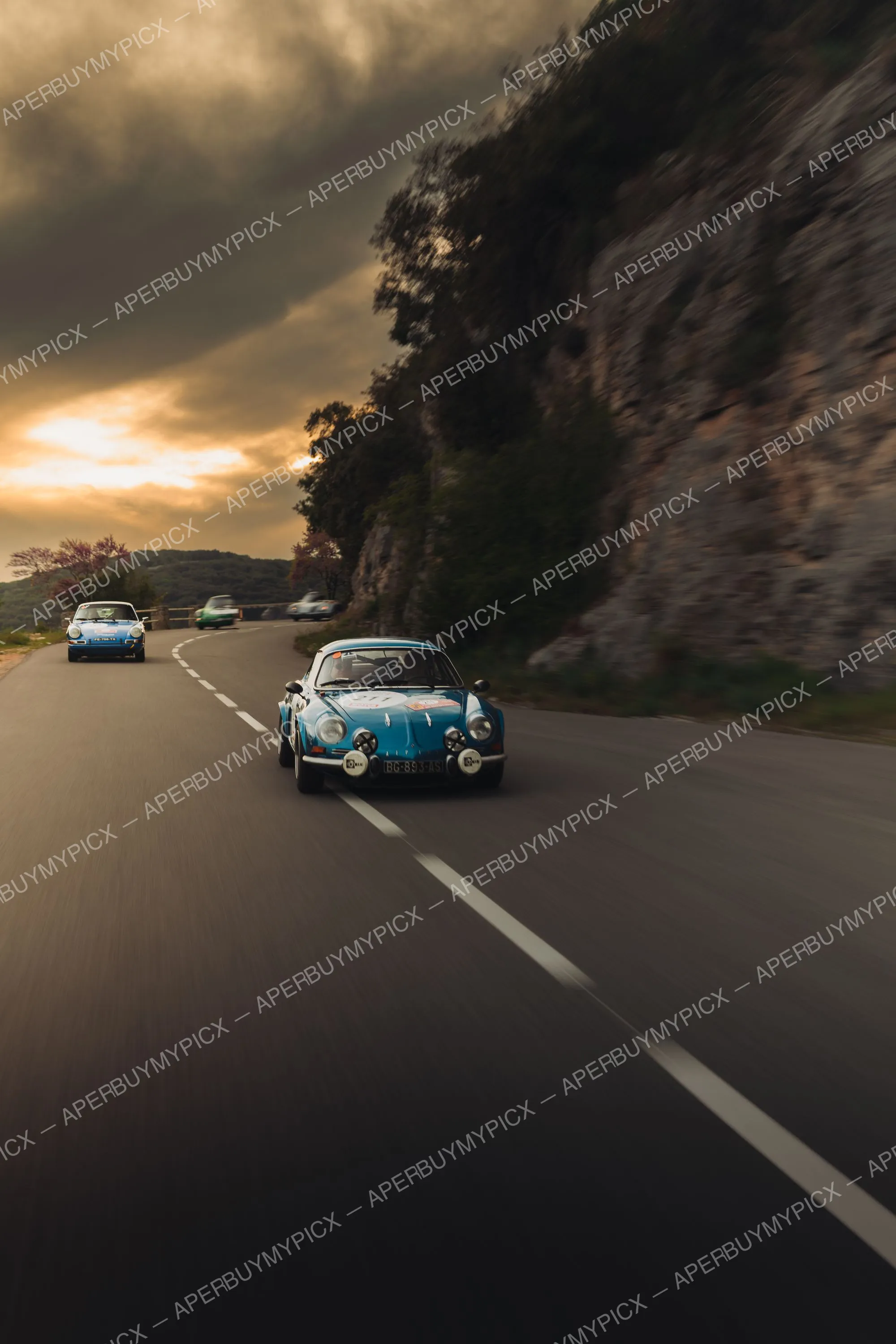 Alpine A110 bleue de rallye roulant sur une route de montagne au coucher du soleil, suivie de Porsche 911 anciennes le long d'une falaise rocheuse, Tour Auto 2025.