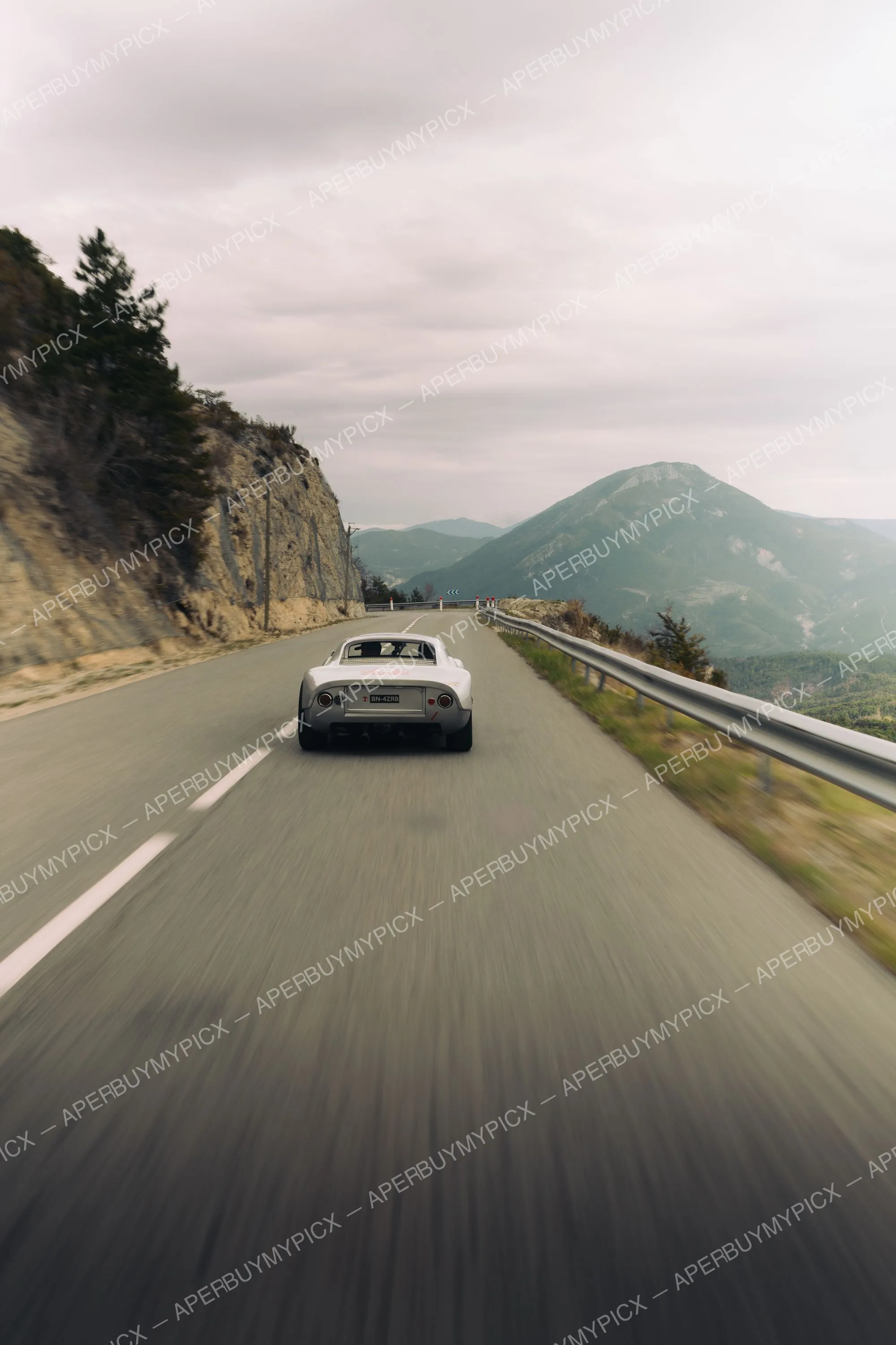 Coupé sportif blanc vu de dos roulant à vive allure sur une route de montagne sinueuse, avec falaise rocheuse, glissière de sécurité et sommets brumeux sous un ciel nuageux.