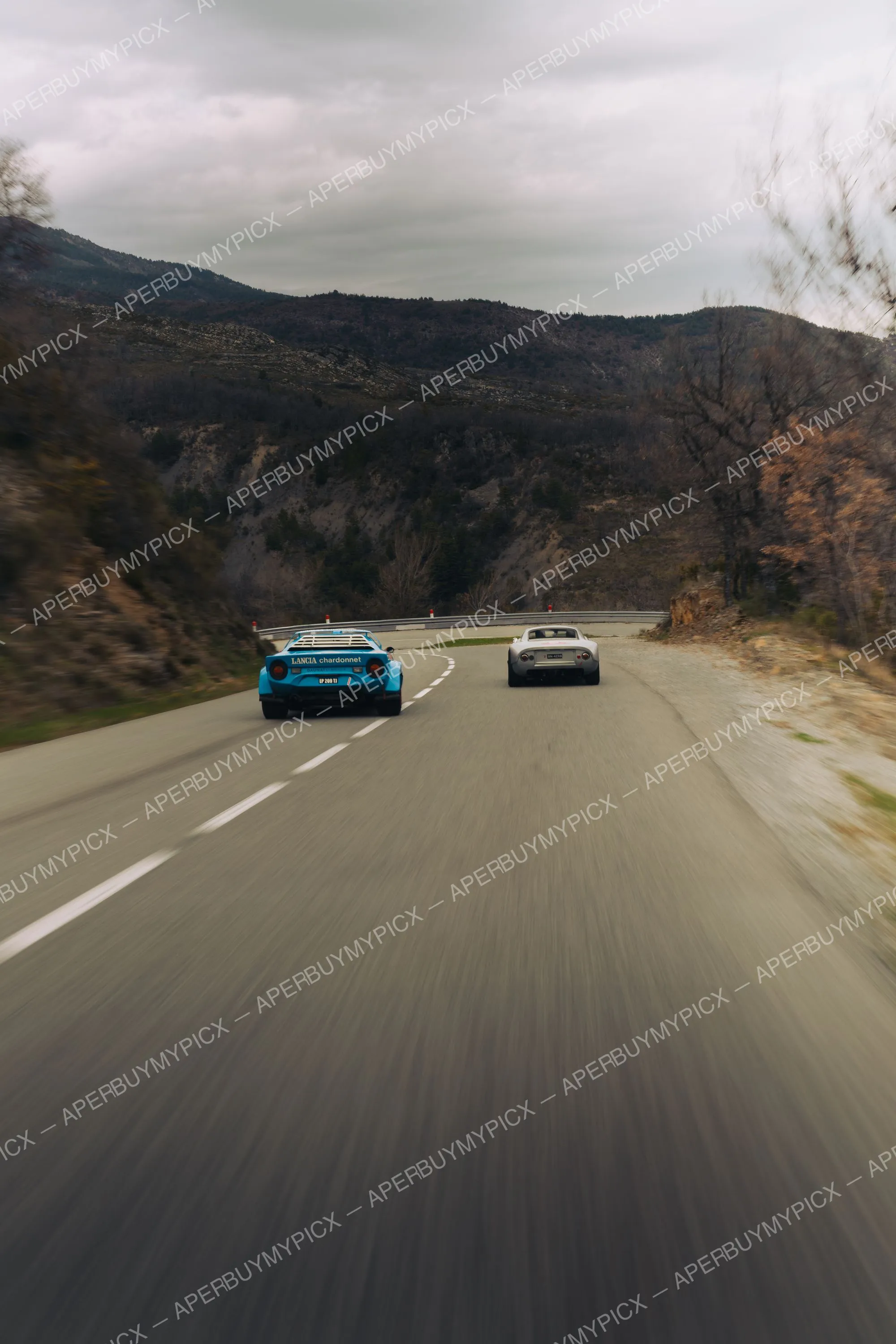 Une Lancia Stratos bleue aux couleurs Chardonnet et une GT argentée roulant à vive allure sur une route sinueuse de montagne sous un ciel nuageux, vue arrière avec effet de vitesse.
