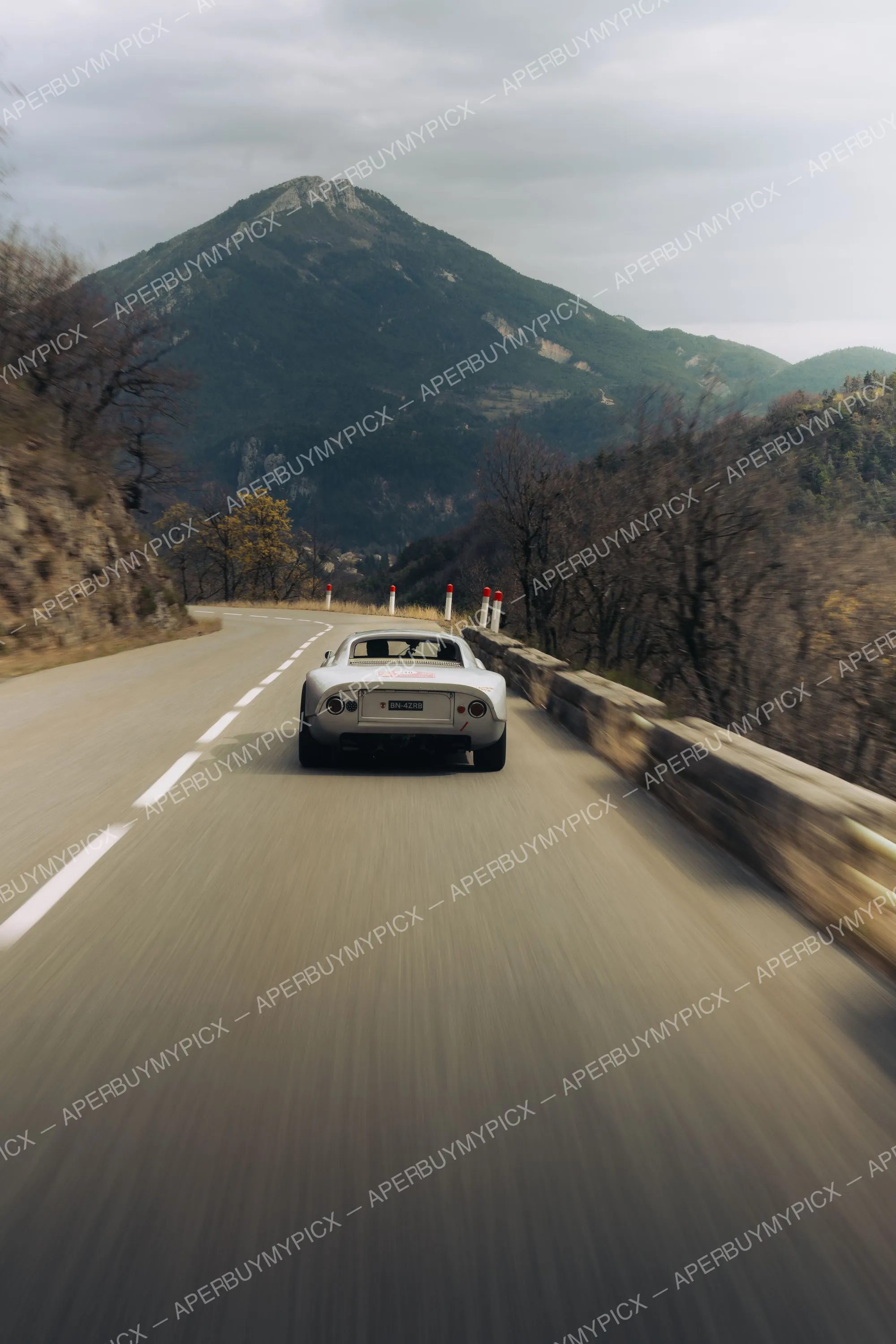 Coupé de sport argenté vu de dos roulant à vive allure sur une route de montagne sinueuse, avec un pic rocheux boisé en arrière-plan et un ciel nuageux